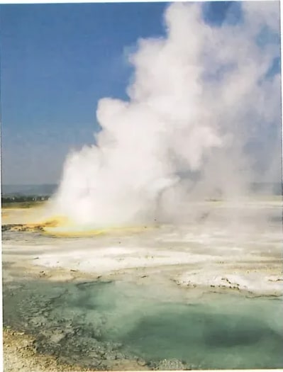 Fotografía a color de un géiser en erupción con columna de vapor blanco sobre un terreno árido y agua turquesa