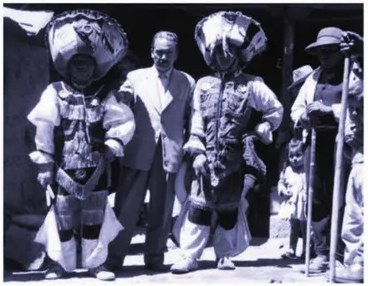 Fotografía en blanco y negro de Arguedas junto a personas con trajes y máscaras de danzantes folclóricos en una festividad.