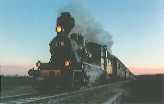 Fotografía de una locomotora de vapor antigua, con el número 1136 visible en su frente, avanzando por las vías con humo blanco saliendo de la chimenea. El fondo muestra un cielo al atardecer con tonos azules y anaranjados. La imagen sirve de estímulo para el ejercicio 10 de escritura de trama descriptiva. Crédito: Shutterstock (2020) 356134373.