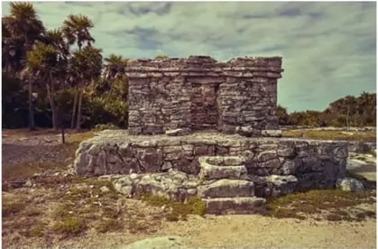 Fotografía de estructura de piedra en un sitio arqueológico con palmeras al fondo.