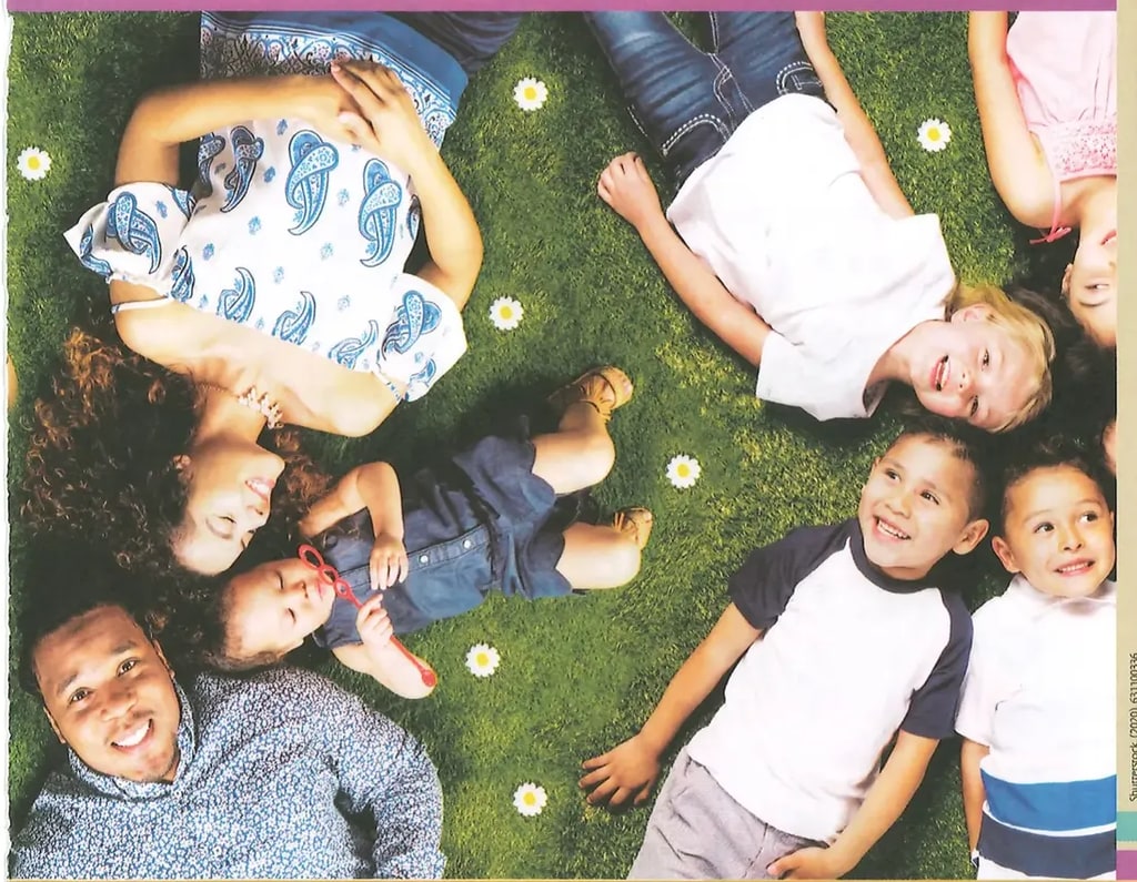 Fotografía de grupo familiar compuesto por un adulto joven (hombre) y varios niños (4-5 niños de distintas edades) acostados en círculo sobre césped verde con flores blancas, todos mirando hacia arriba (hacia la cámara). La imagen transmite diversidad, unión familiar y alegría. Ocupa aproximadamente la mitad central de la página. Crédito: Shutterstock (2020) 631100336.