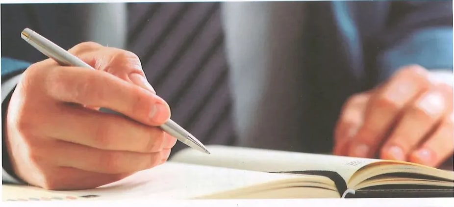 Fotografía a color de manos sosteniendo un bolígrafo plateado escribiendo en un libro abierto
