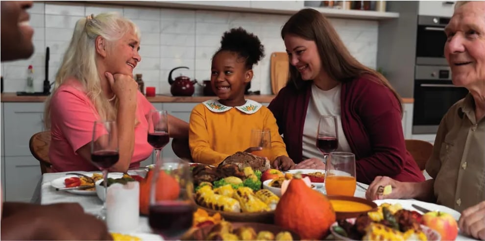 Fotografía de una familia multigeneracional reunida alrededor de una mesa con un pavo asado, mazorcas de maíz, frutas y vino; representa una cena típica de Thanksgiving.