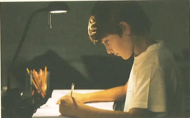 Fotografía de un joven estudiante de perfil escribiendo en un cuaderno bajo una lámpara de escritorio, en un ambiente de estudio nocturno. Imagen que acompaña la sección de gramática. Crédito: Shutterstock (2020) 290611184.