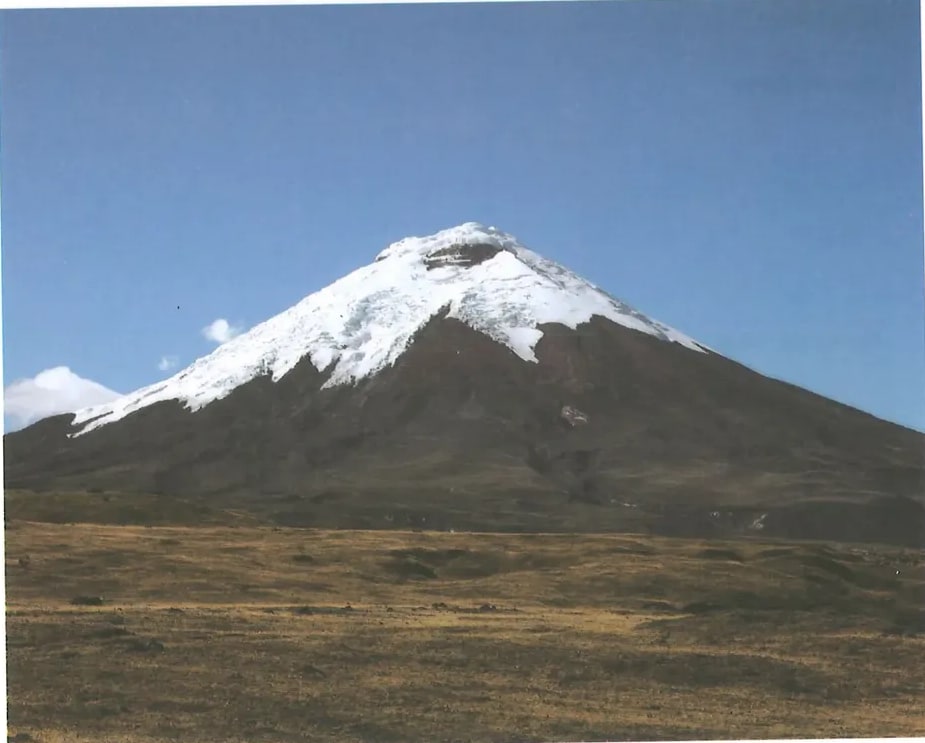Fotografía grande de un volcán nevado en erupción o con penacho de humo/vapor, con nieve en la cumbre y ladera. El cielo es azul con algunas nubes. Probablemente el volcán Cotopaxi (Ecuador), uno de los volcanes más altos del mundo con cumbre nevada. La fotografía ilustra el tema del texto expositivo sobre la actividad volcánica. Ocupa la mitad inferior de la página. Crédito: Shutterstock (2020) 240040720.