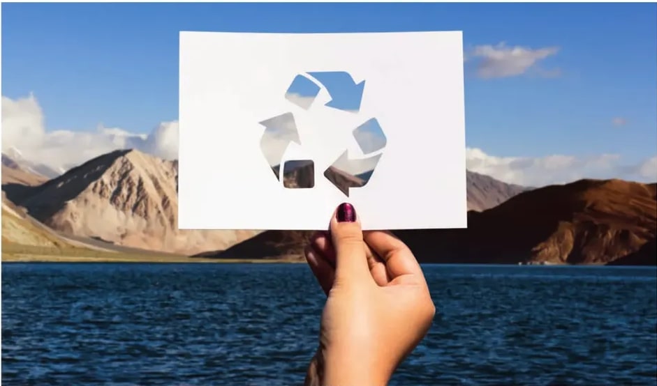 Fotografía de una mano sosteniendo un papel blanco con el símbolo de reciclaje recortado, sobre un fondo de montañas y un lago azul intenso.