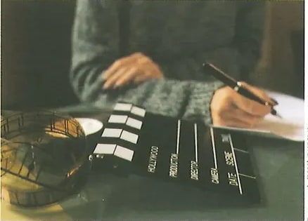 Fotografía de manos de una persona escribiendo en papel con bolígrafo. Al fondo se ven clappers (claquetas) de filmación cinematográfica, evocando el contexto de la reseña de películas. Crédito: Shutterstock (2020) 540260491.